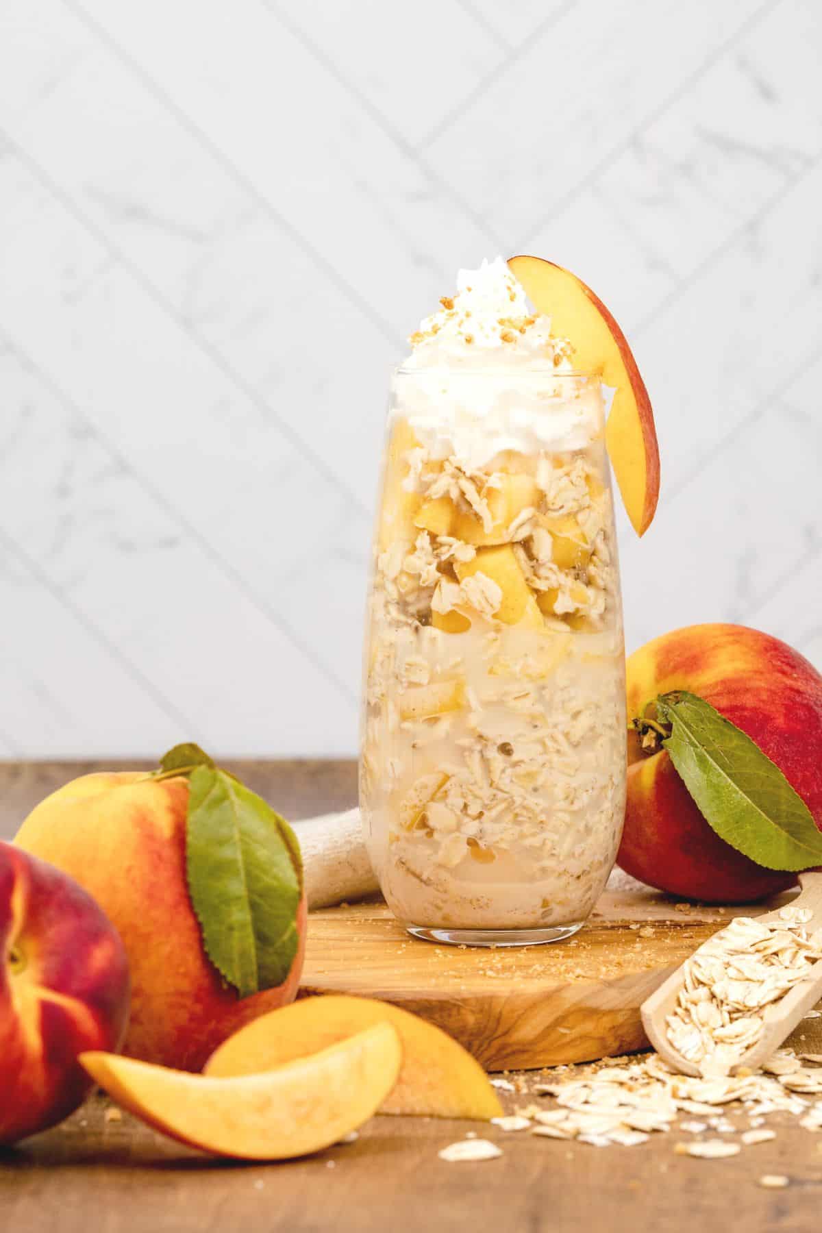peach overnight oats in a glass flute cup with whip cream on top. peaches and slices of peaches surround the glass with some raw oats.