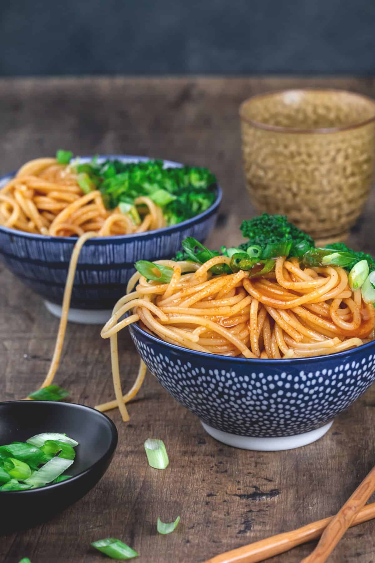 two blue bowls seen from the side filled overflowing with gochujang noodles and broccoli. a cup and chopsticks are also seen.