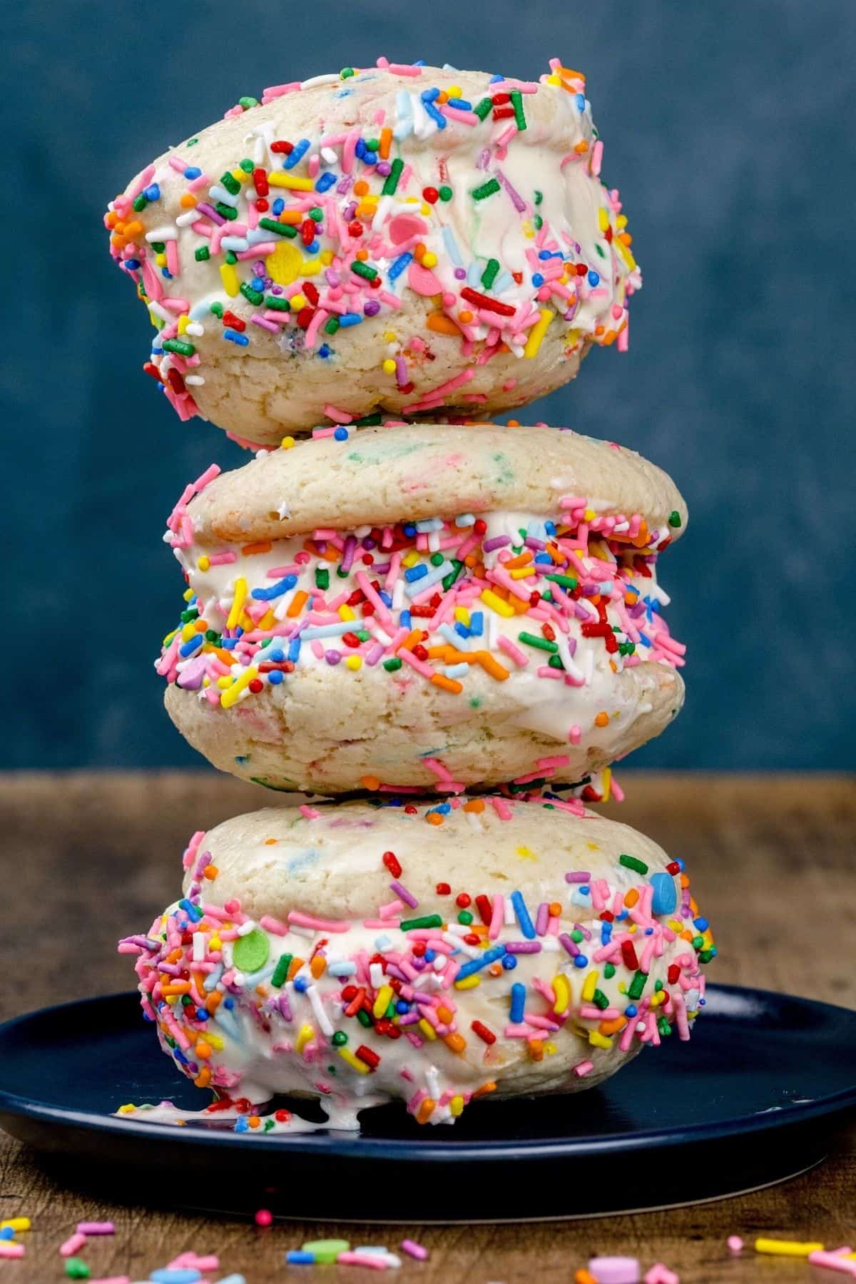a stack of 3 rainbow sprinkled gluten free ice cream sandwiches on a dark blue plate in front of a dark blue background