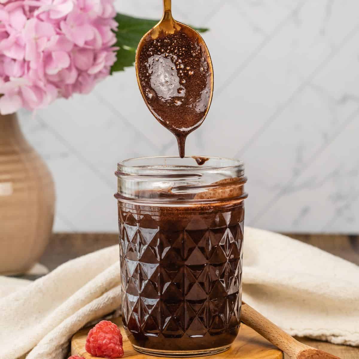 mason jar filled with vegan chocolate sauce with a spoon pouring more into the jar. a vase of flowers is blurred in the background