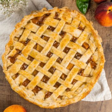 peach pie with a lattice crust is shown on a wood table with fresh peaches and herbs surrounding it