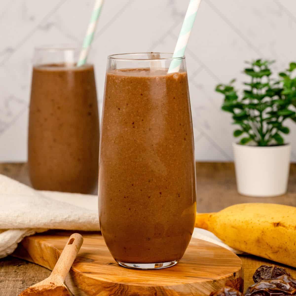 two tall glasses are filled with a chocolate dairy free weight gain shake. there are blue and white straws in each glass. they are sitting on a wood table top surrounded by cocoa powder, bananas, and more fresh dates.