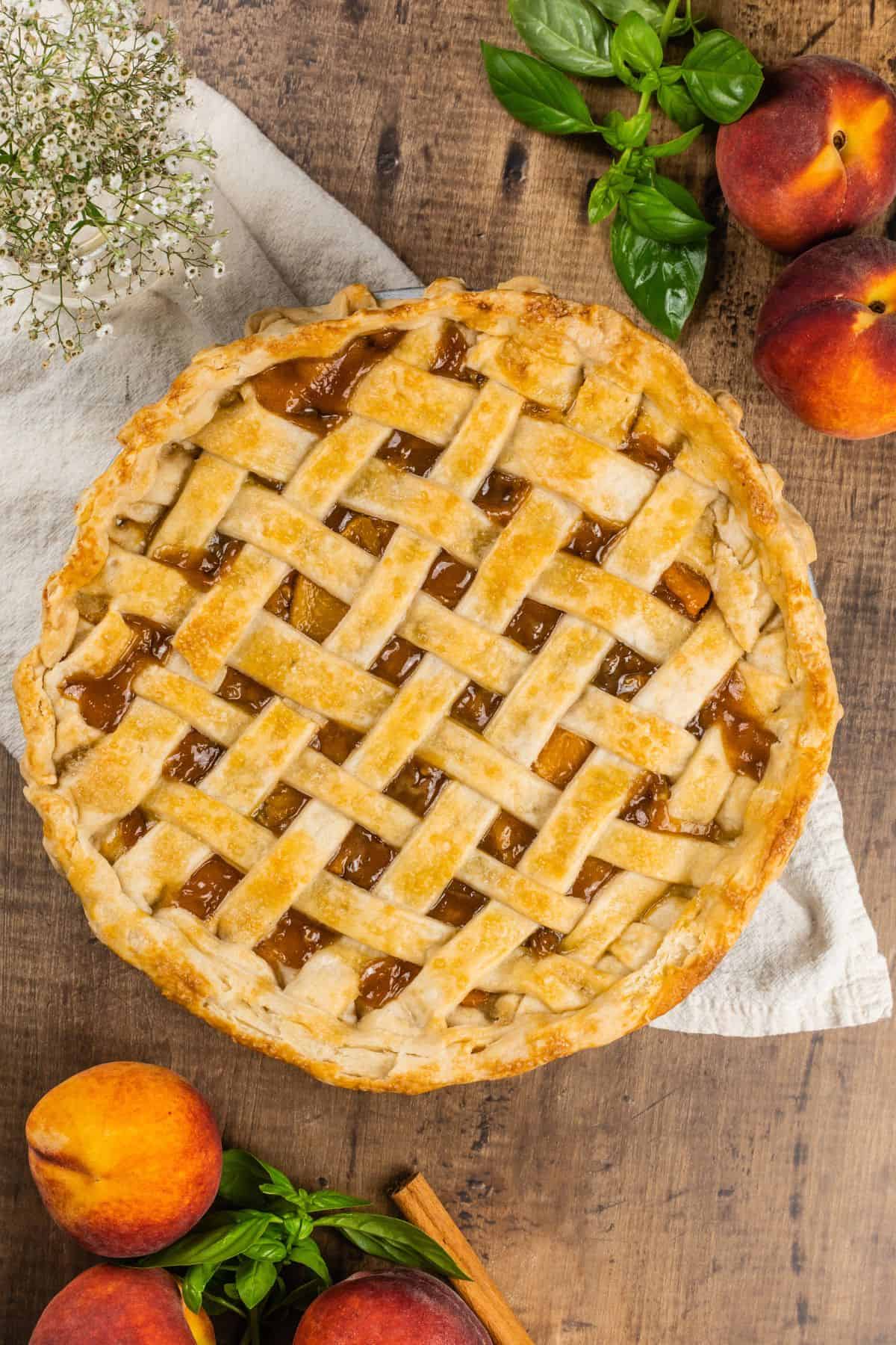 finished peach pie with a lattice crust surrounded by peaches and herbs on a wood table