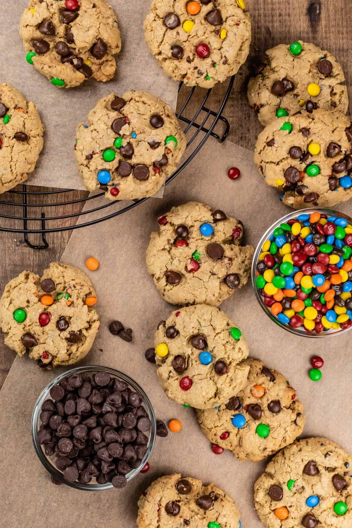 many gluten free monster cookies on the wood table with parchment paper and bowls of chocolate chips and m&ms.