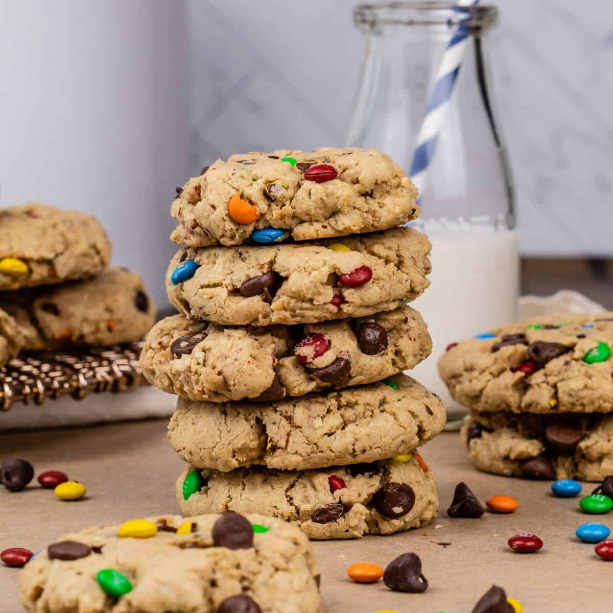 a stack of 5 gluten free monster cookies on a table surrounded by more cookies, chocolate chips, candies, and a glass of milk blurred in the background.