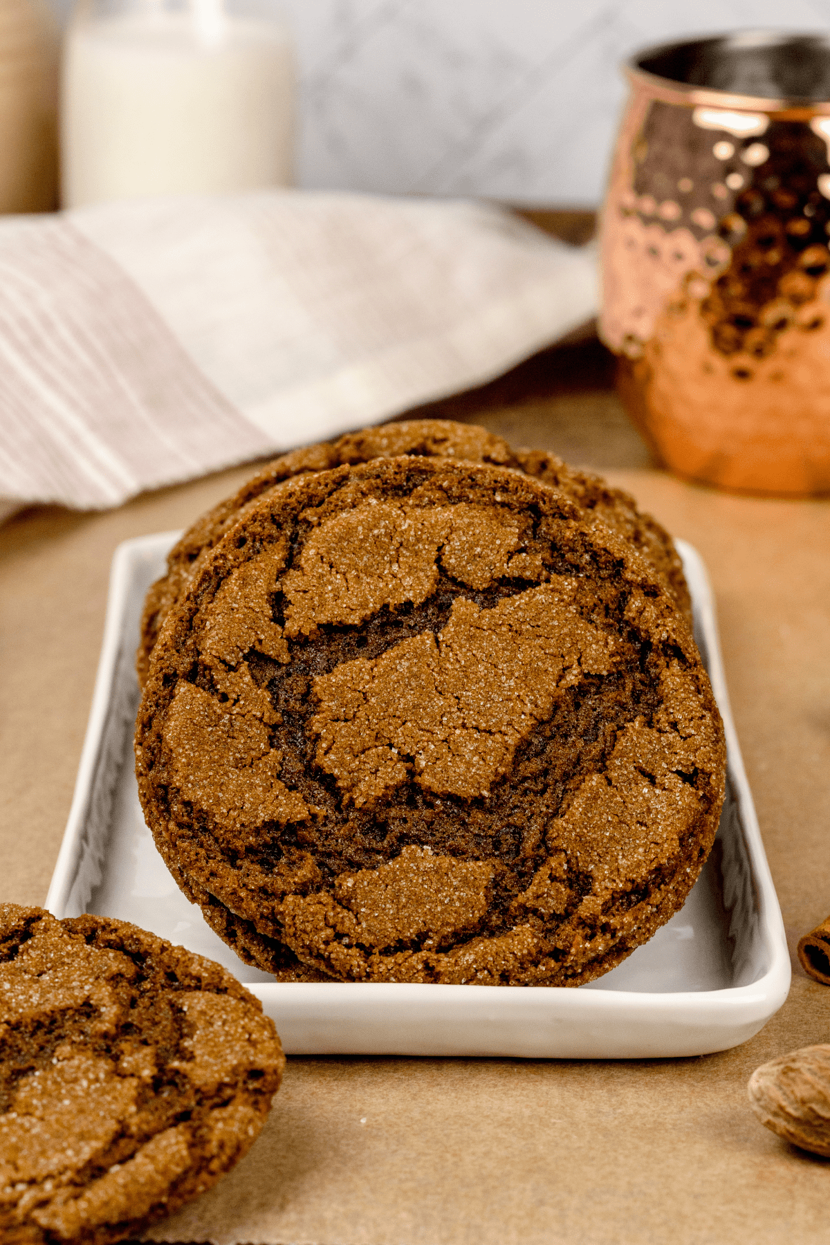 a white plate filled with vegan ginger cookies up close so you can see the crinkle texture of the top of the cookie. milk glasses and a towel are blurred in the background.