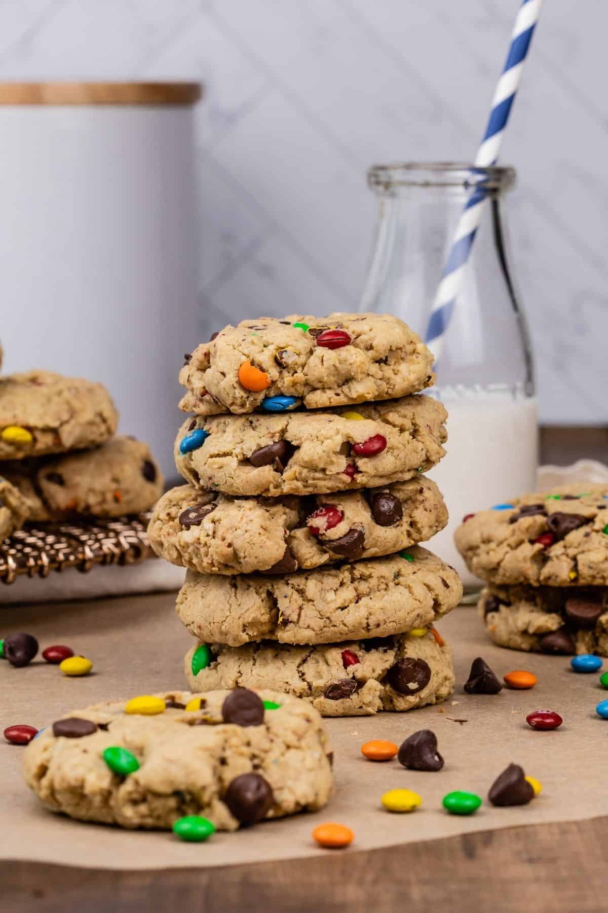 a stack of 5 gluten free monster cookies on parchment paper. more cookies, chocolate chips, and m&ms surround the cookies. a glass of milk with a straw is blurred in the background.