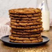 stack of many vegan ginger cookies on a dark blue plate with sugar sprinkled around and a glass of milk blurred in the background.