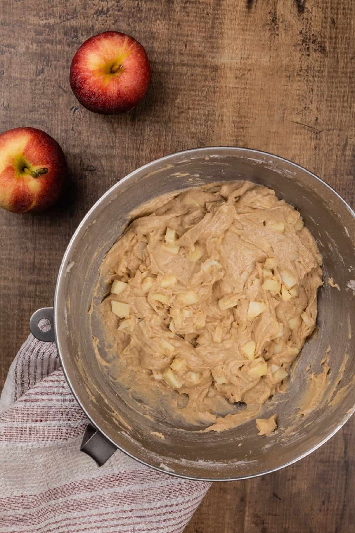 a silver bowl filled with the finished apple cinnamon muffin batter and you can see the apple chunks inside the batter. a red and white stripe towel is under the bowl. two apples are next to the bowl.