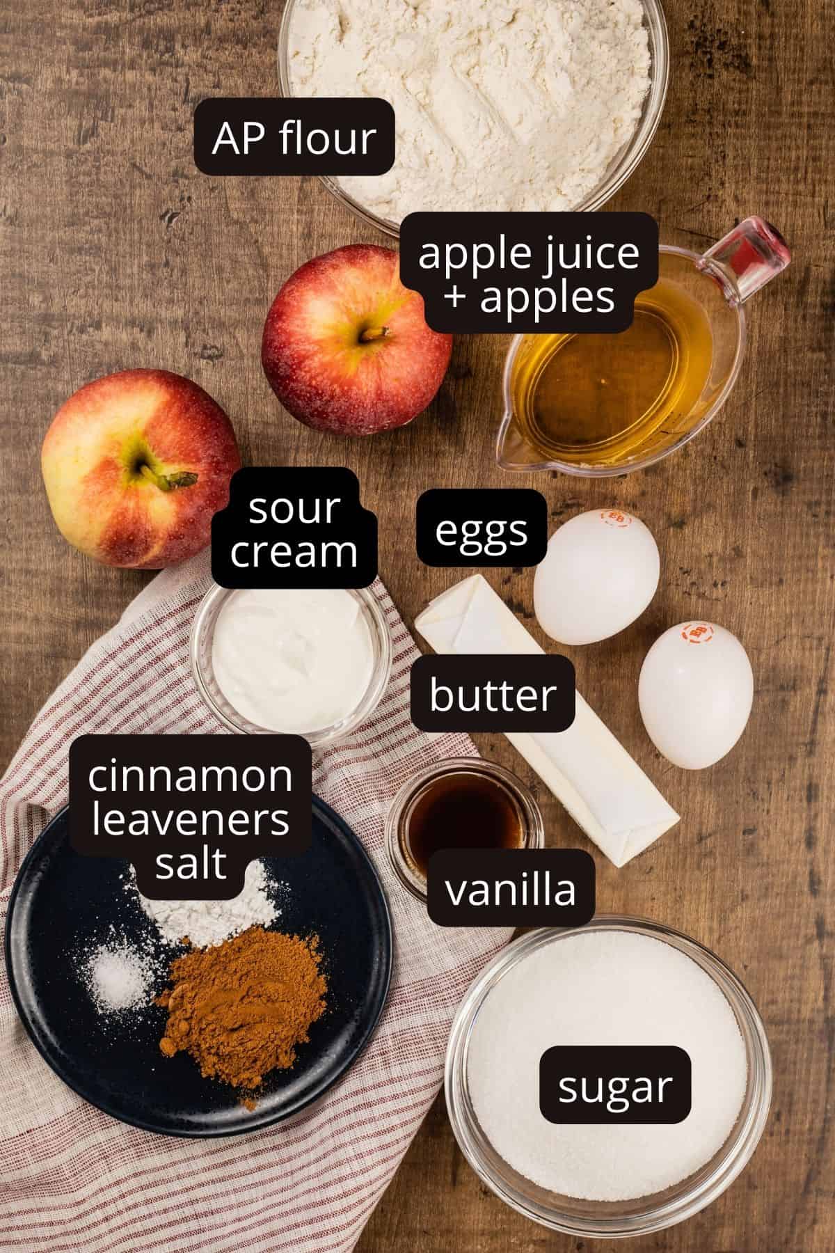 ingredients to apple cinnamon muffins in various glass bowls on the wood tabletop. black text boxes with white lettering labels each ingredient.