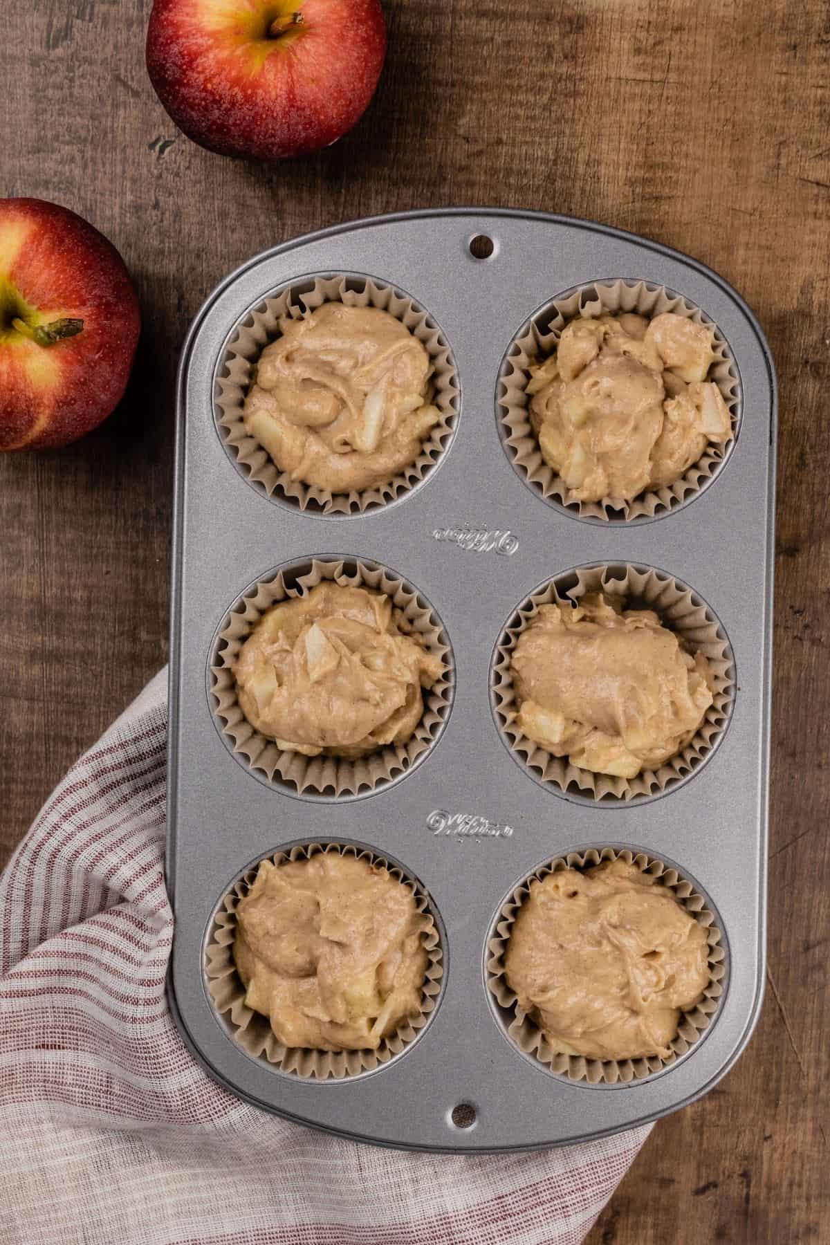 filled 6 muffin tray with the raw apple cinnamon muffin batter. a red and white stripe towel is under the tray. two apples are next to the tray.