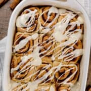 cinnamon rolls in a white baking tray covered with icing. a hint of a towel and cinnamon sticks can be seen on the table as well.