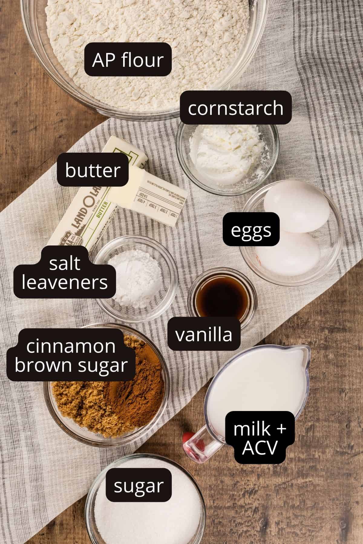 ingredients for cinnamon rolls without yeast in various glass bowls on a wood tabletop. a white and gray towel is also seen. black and white labels have been added to identify each ingredient.