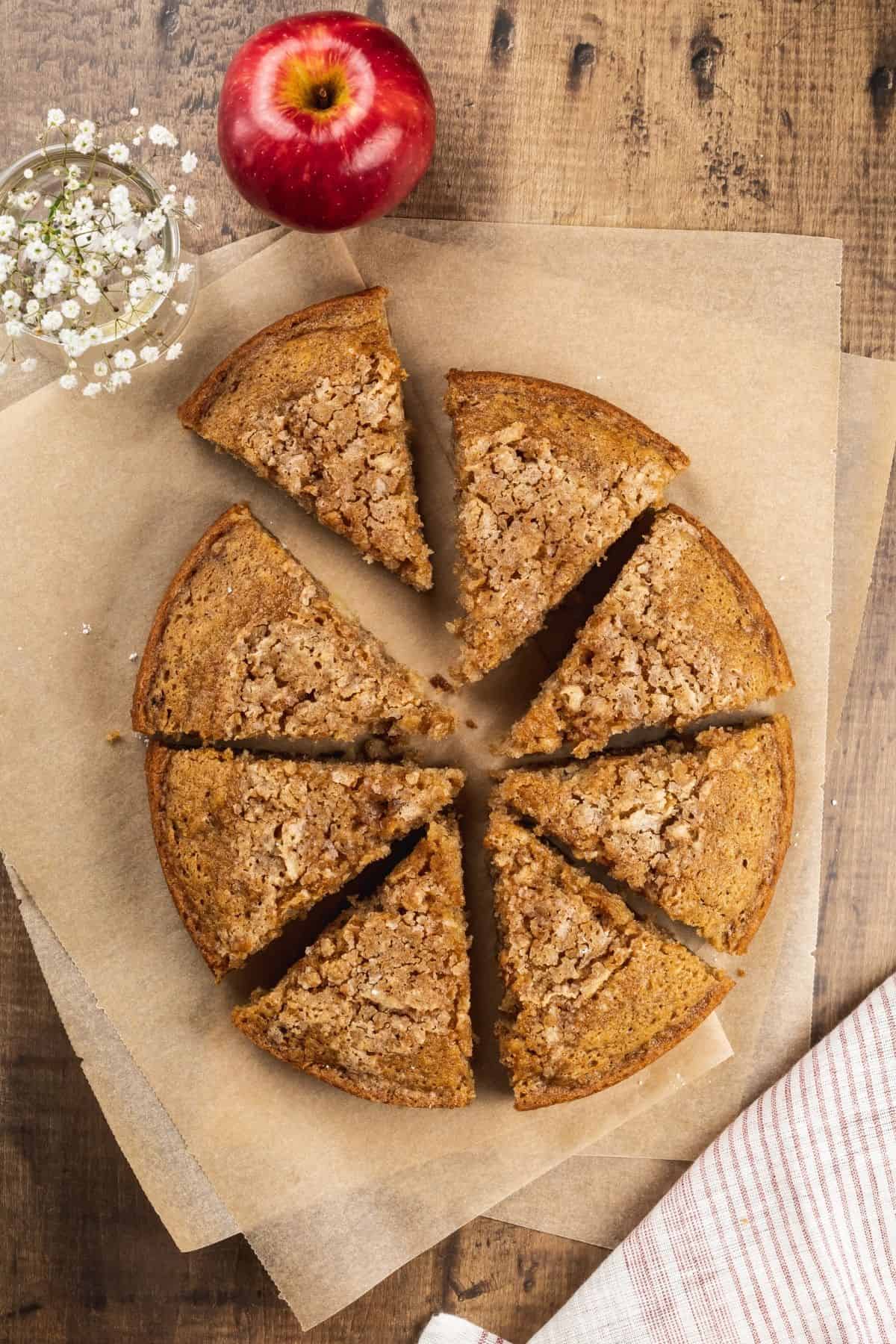 triangle slices of a round vegan apple cake are on brown parchment paper with tiny flowers in a vase next to it as well as an apple.