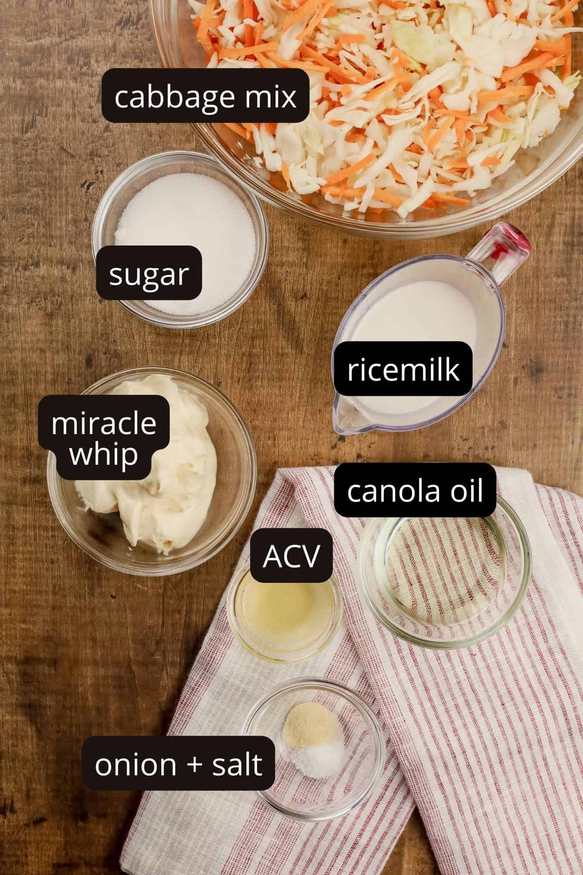 ingredients for dairy free coleslaw on  a wood table in various glass bowls. black and white labels have been added to each ingredient. 
