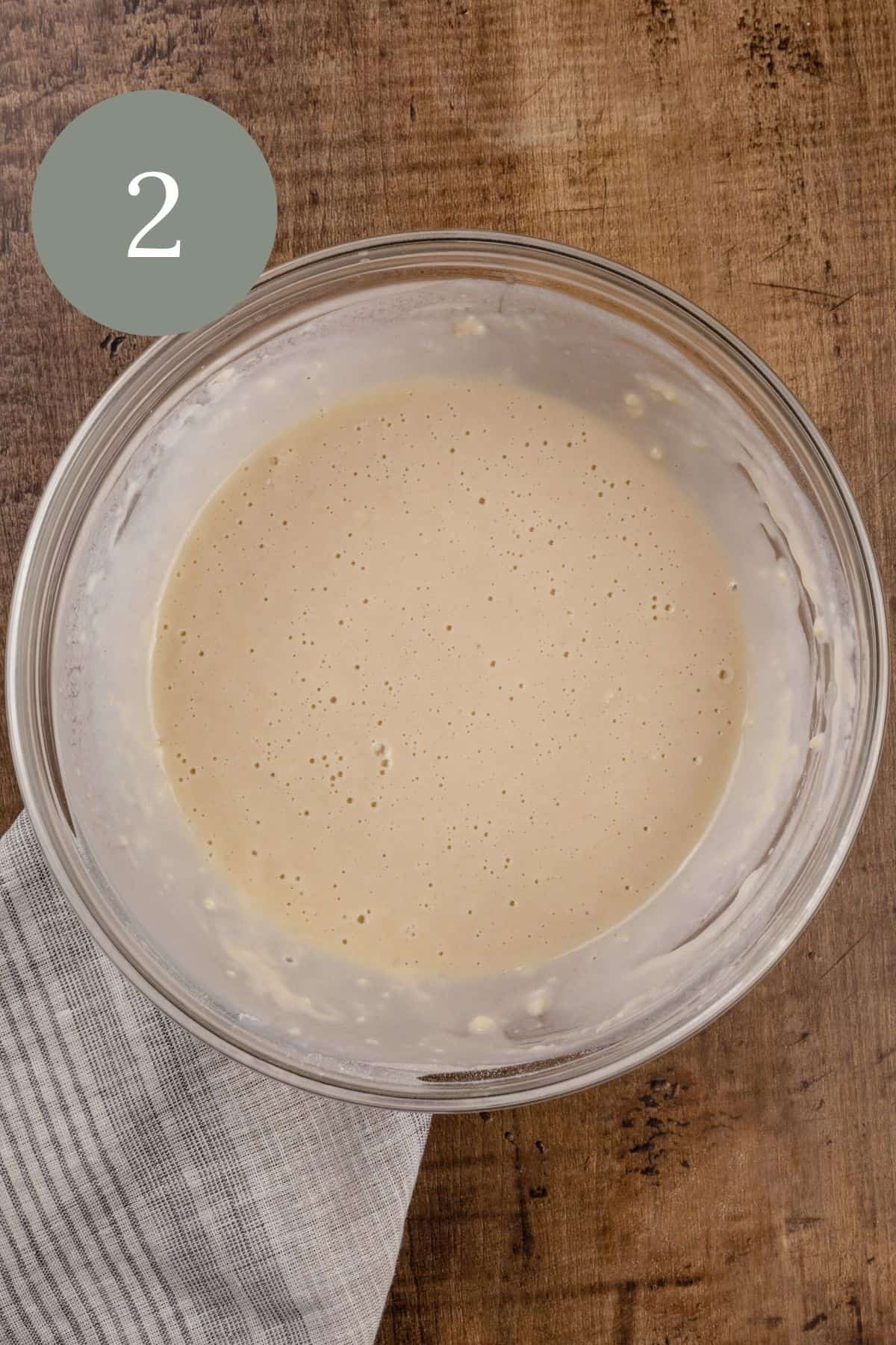 the finished pancake batter in a glass bowl on the wood tabletop. a gray and white stripe towel is under the bowl. a round green circle with the number two is in the top left corner.