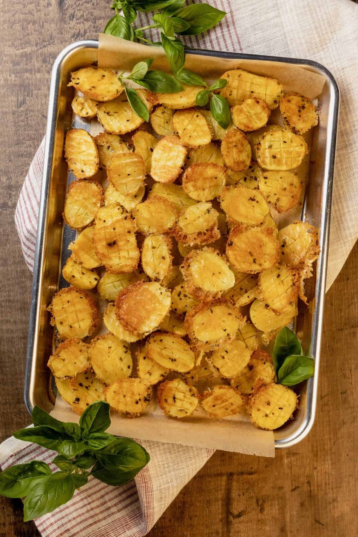 parmesan potatoes with fresh basil on a baking sheet on a wood table. a red and white towel is under the baking sheet.