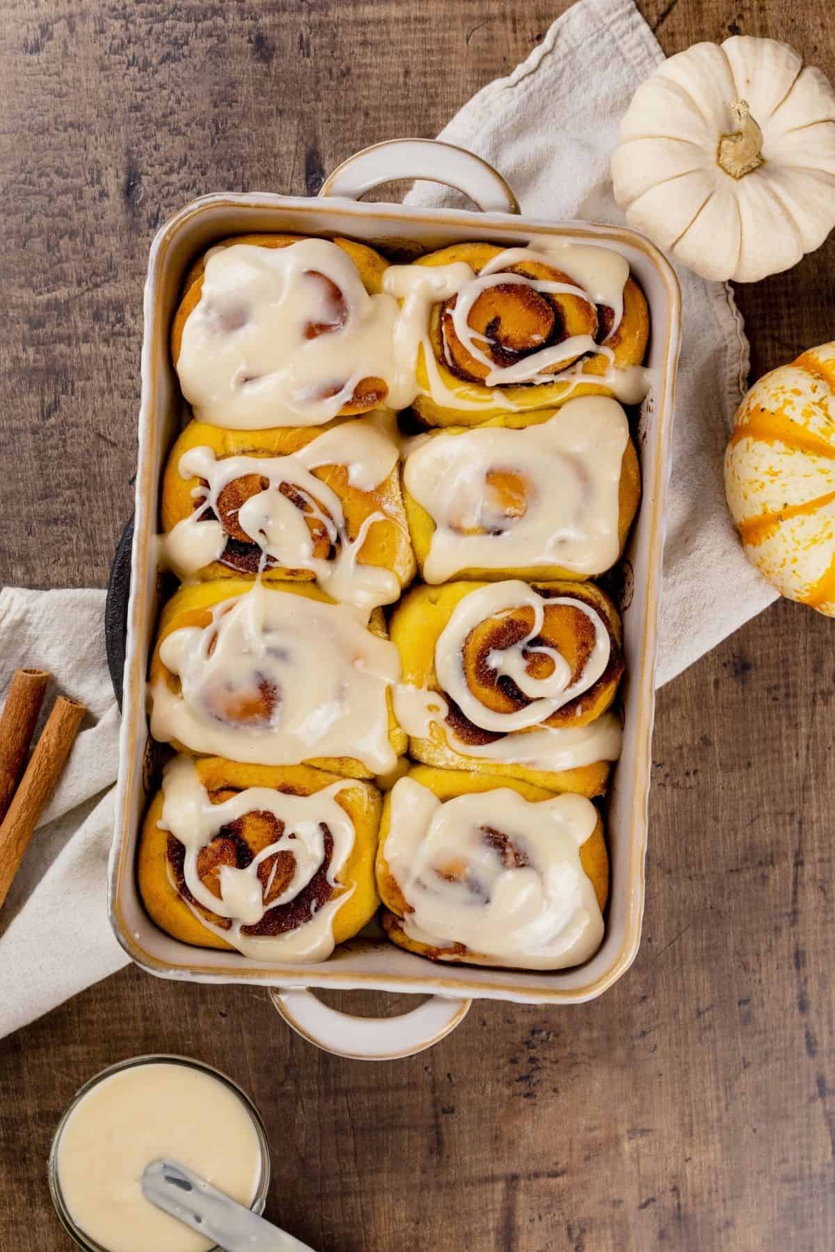 looking down on a baking dish filled with 8 pumpkin cinnamon rolls covered in icing. the dish is on a wood table. mini pumpkins, cinnamon sticks, and a bowl of extra icing is next to the dish.