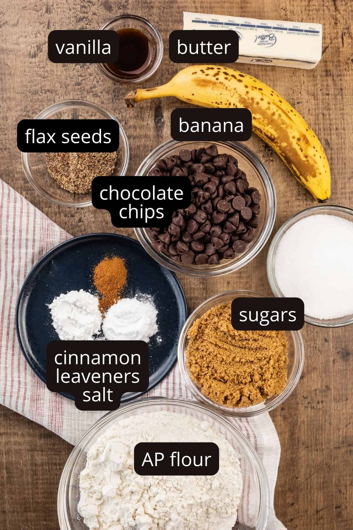 ingredients for vegan banana chocolate chip cookies in various glass bowls on the wood tabletop. a red and white strip towel in under some of the bowls. black and white labels tell you what the ingredients are.