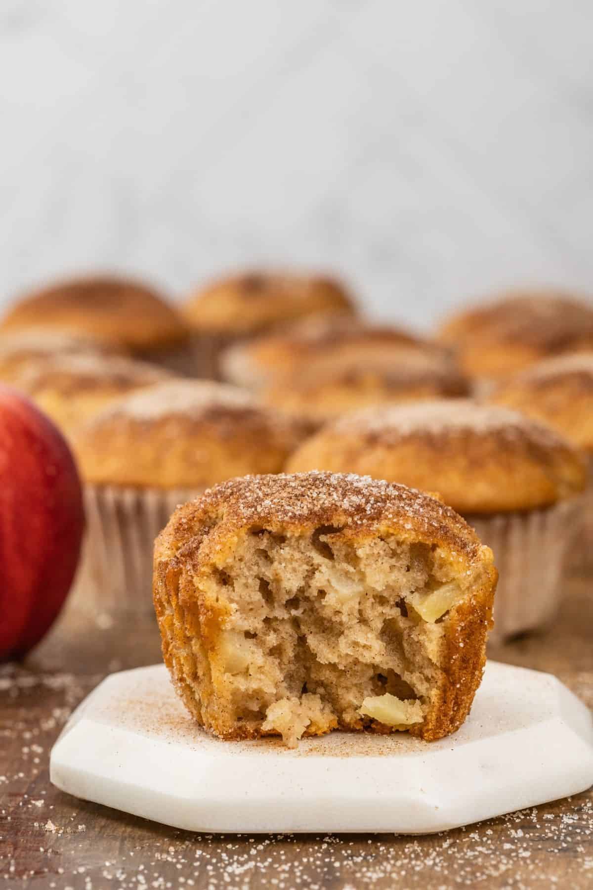 a single apple cinnamon muffin rests on a marble coaster. a bite is taken out of the muffin to show off the texture on the inside. more muffins and apples are blurred in the background.