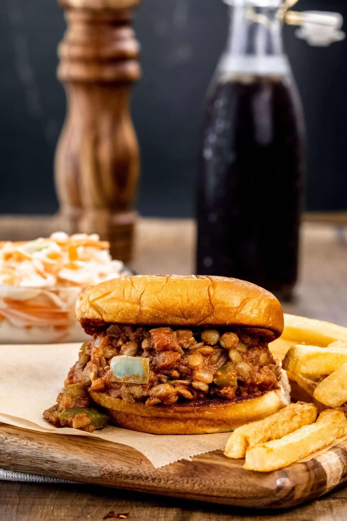 a lentil sloppy Joe on a toasted bun next to a pile of French fries. it is on a serving board. a bowl of coleslaw and a bottle of soda are blurred in the background.