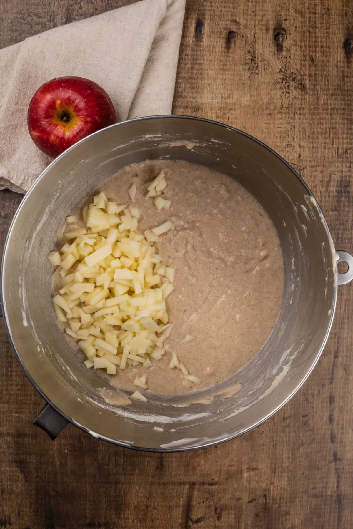adding diced apple chunks to the apple cake batter in a silver mixing bowl on a wood table with an apple next to the bowl.