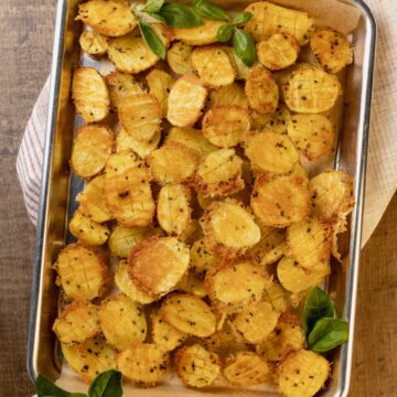 parmesan potatoes with fresh basil on a baking sheet. a red and white towel is under the baking sheet.