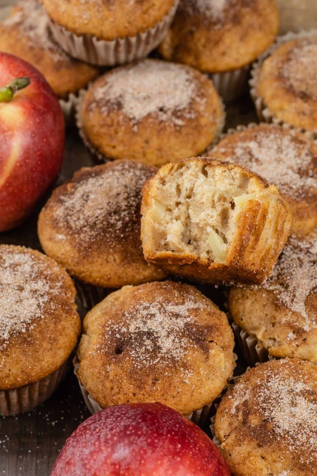 a pile of apple cinnamon muffins on the table with apples mixed in. one muffin is on top of the others and a bite is taken out of it.
