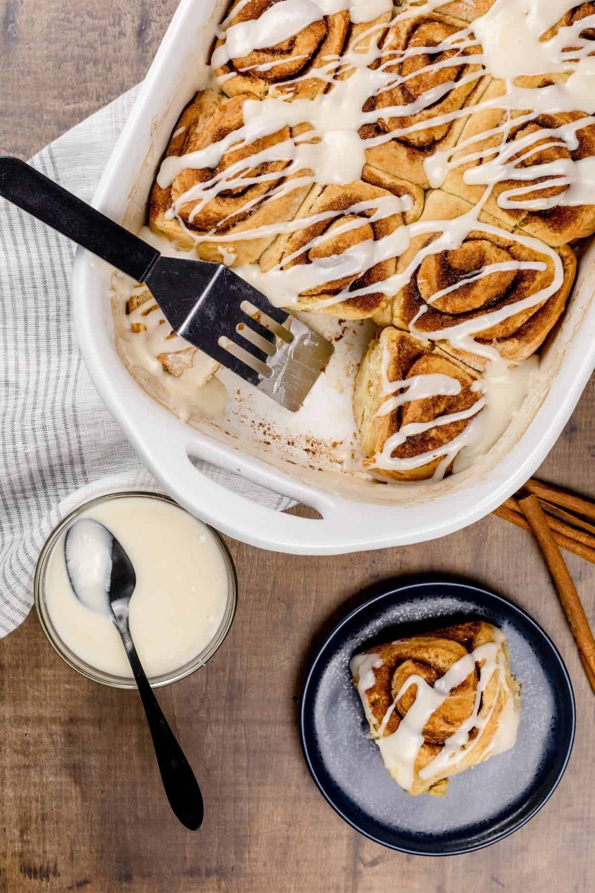 fully baked cinnamon rolls being served with a silver serving spoon. a roll is on a blue plate. the bowl of icing with a spoon is next to the plate.