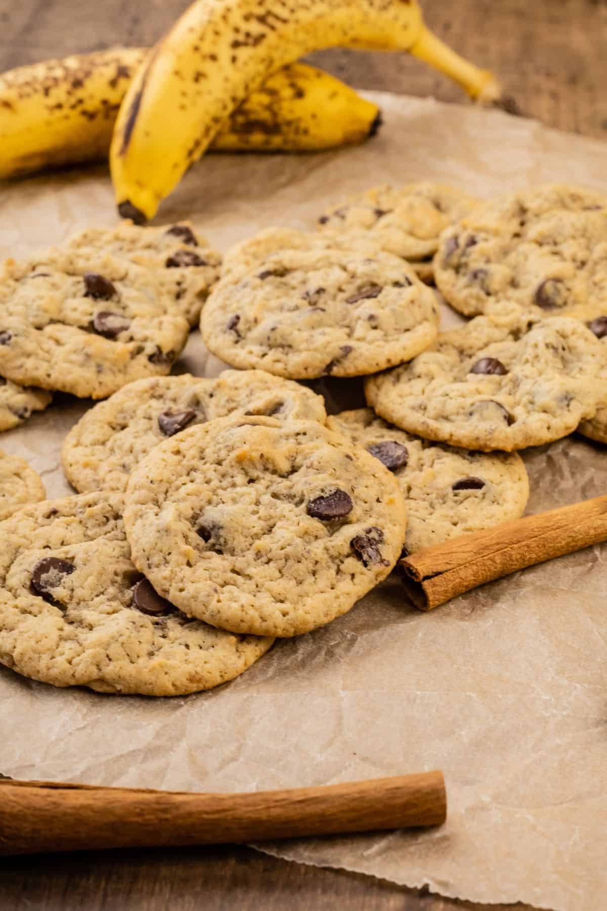 side view of vegan banana chocolate chip cookies laying in a pile on top of crinkled brown parchment paper. a cinnamon stick is blurred in the foreground and two bananas are blurred in the background.