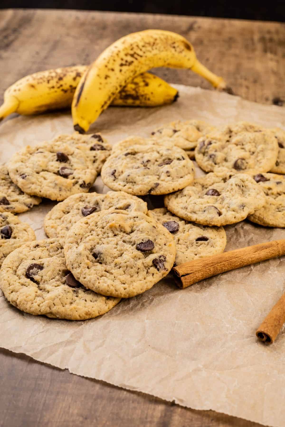 a side view of a pile of many vegan banana chocolate chip cookies with cinnamon sticks and bananas blurred in the background. it is resting on a wood tabletop and you can see just the edge of the table.