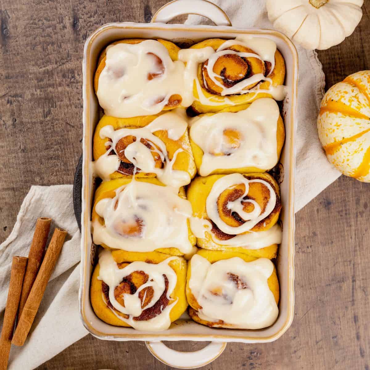 looking down on 8 pumpkin cinnamon rolls with icing on top. small pumpkins and cinnamon sticks are next to the baking dish.