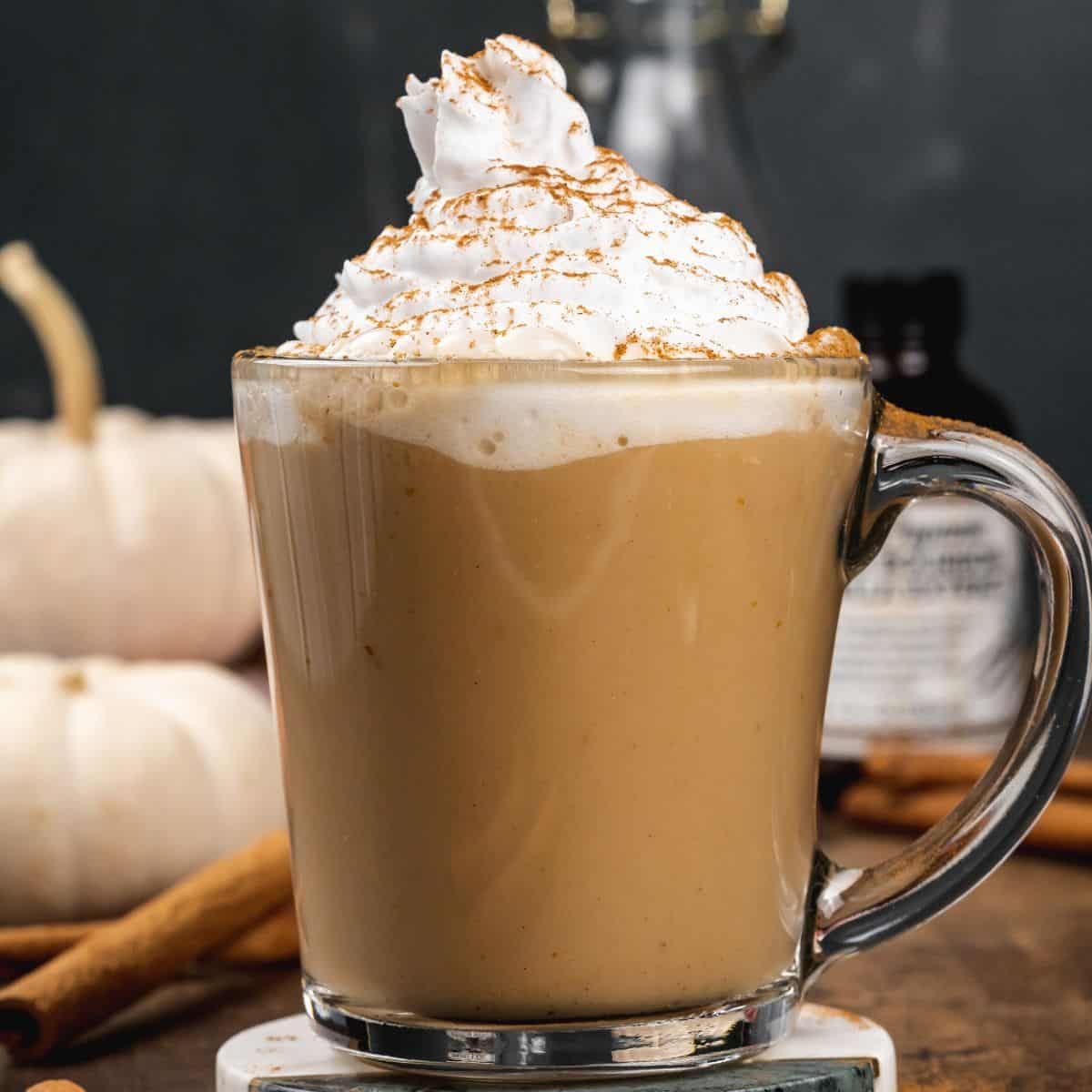 a glass mug filled with pumpkin spice latte with lots of coconut whip cream on top and is sprinkled with cinnamon. it is in front of mini pumpkins and cinnamon sticks blurred in front of a dark background.