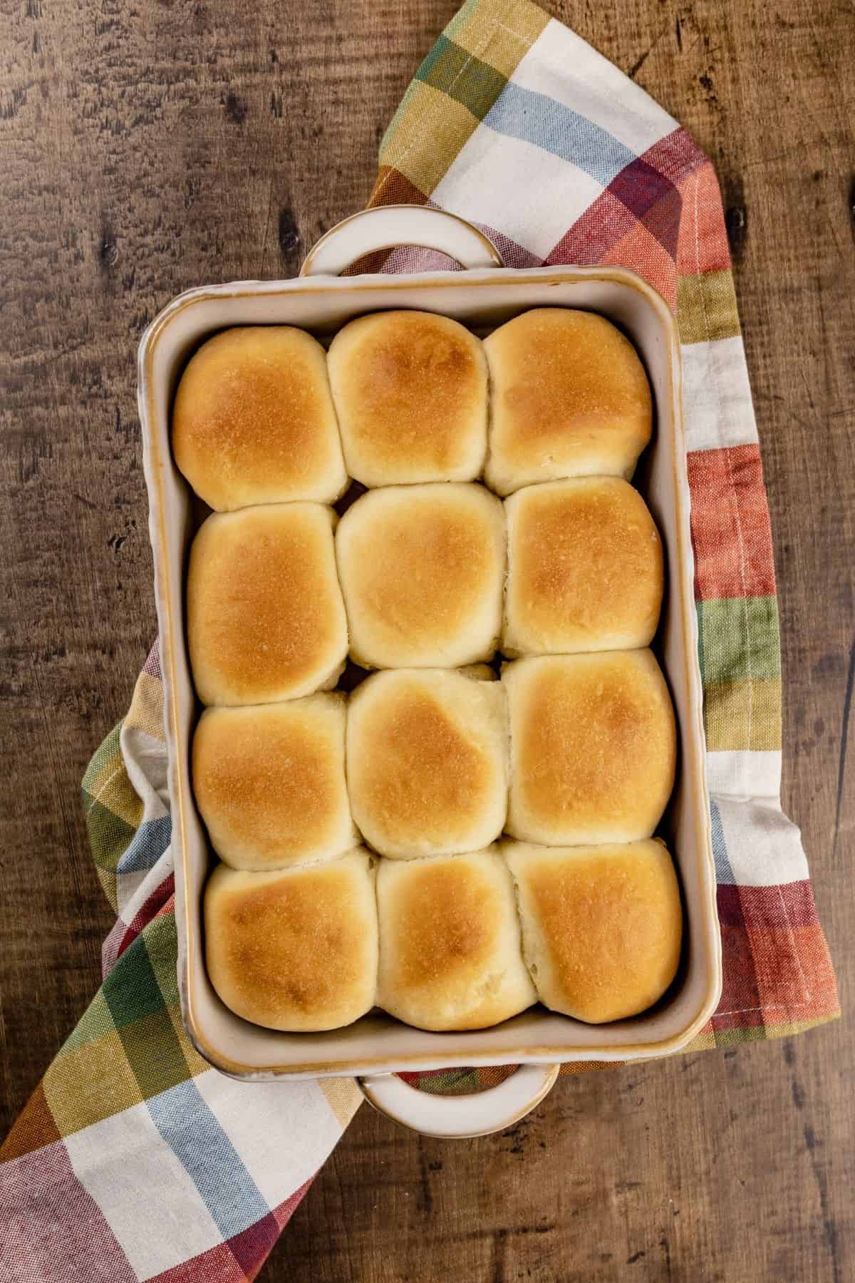 12 baked rolls in a baking dish on top of a kitchen towel on the kitchen wood tabletop.