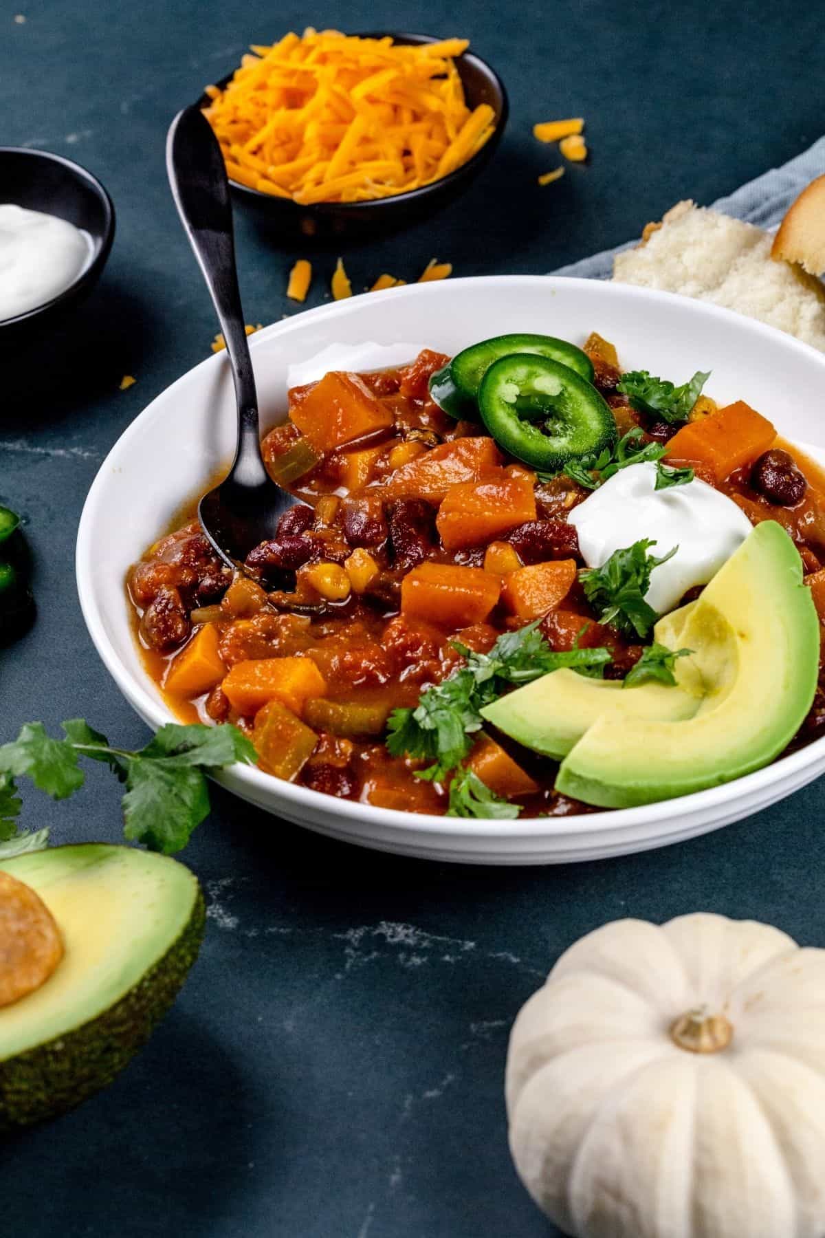 side view of a white bowl filled with pumpkin chili and various chili toppings like avocado and sour cream. a mini white pumpkin is blurred in the foreground.