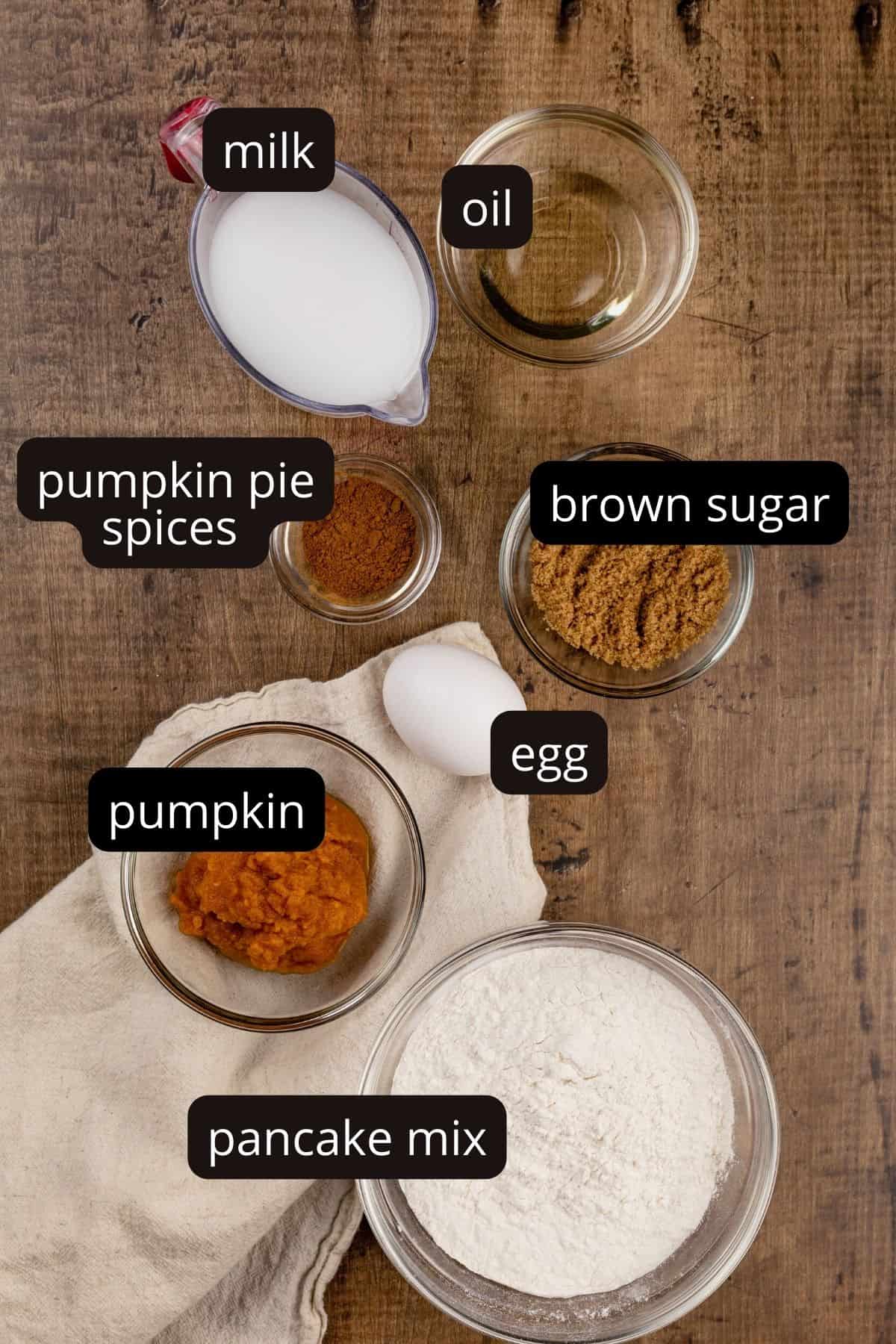 ingredients for pumpkin pancakes in various glass bowls on a wood tabletop. black and white labels have been added to show each ingredient.