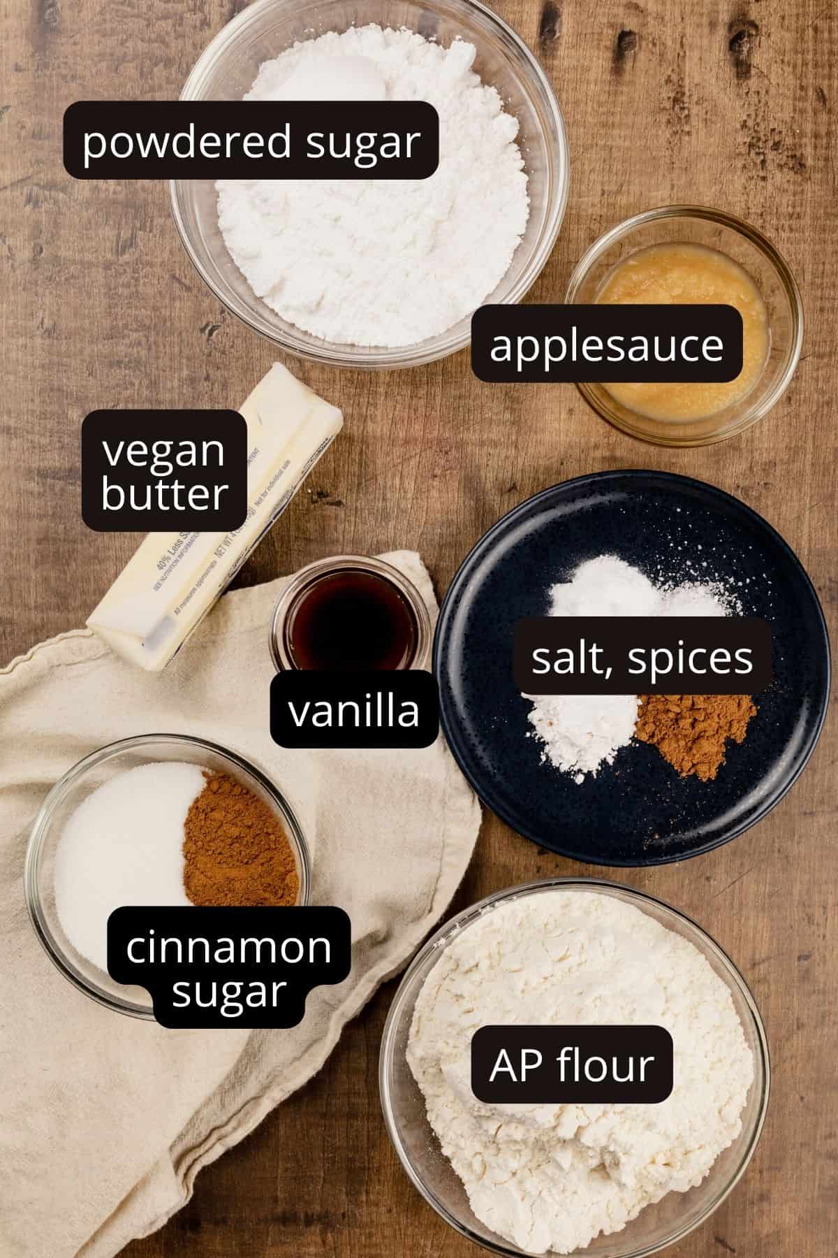 ingredients for vegan snickerdoodle cookies in various glass bowls on a wood table top. black and white labels have been added to name each ingredient.