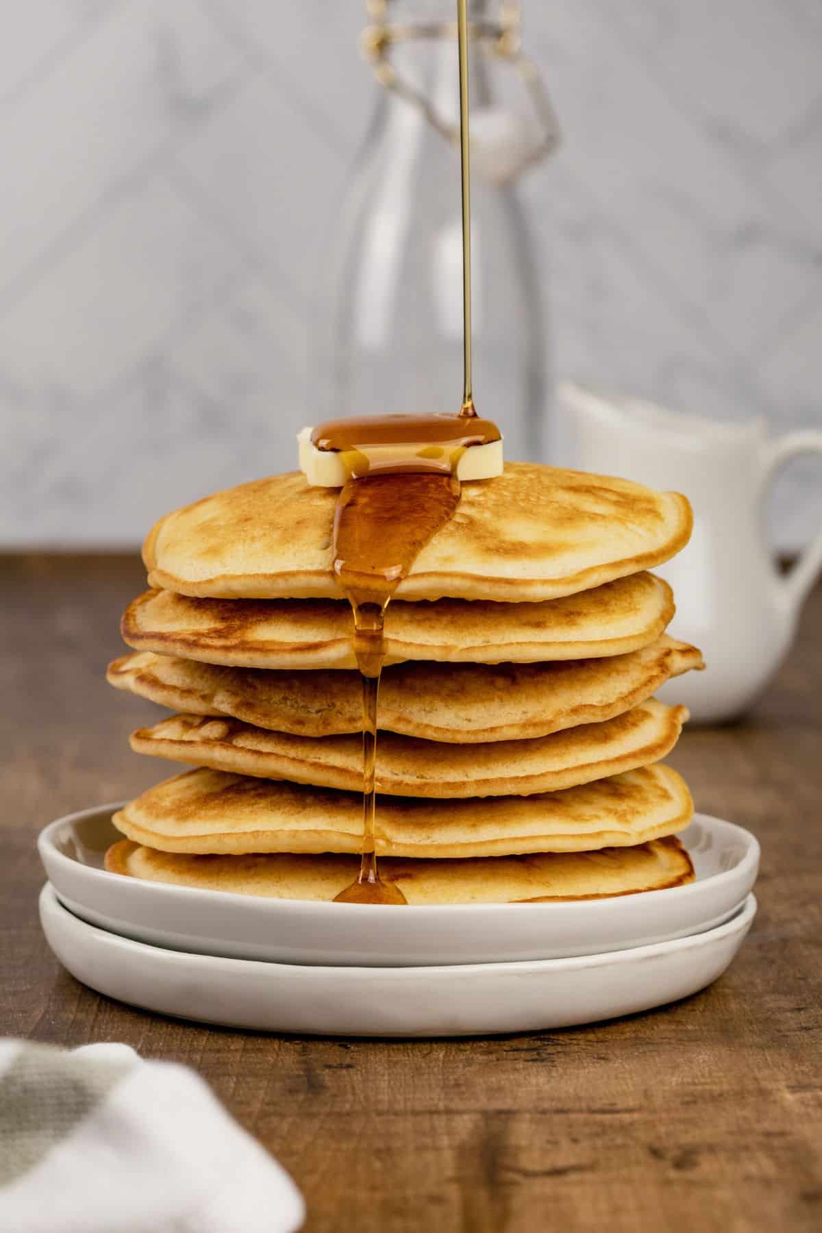 stack of pancakes on a white plate with syrup being poured on top and is dripping over the sides of the pancakes.