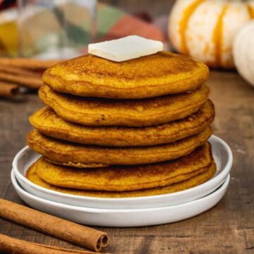a stack of pumpkin pancakes on a white plate with a pat of butter on top. cinnamon sticks and mini pumpkins are blurred in the background.