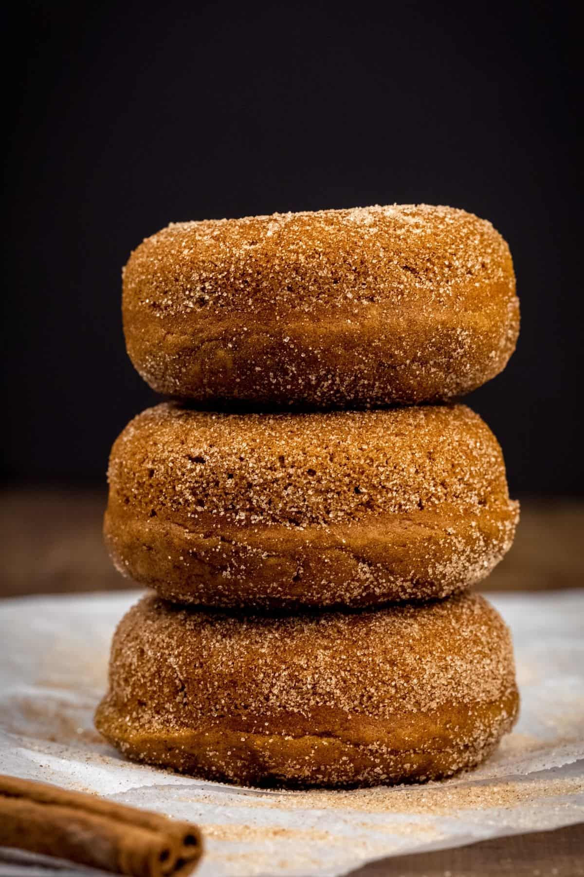 a stack of 3 donuts on top of each other on white parchment paper in front of a black background. a single cinnamon stick is next to the donuts.