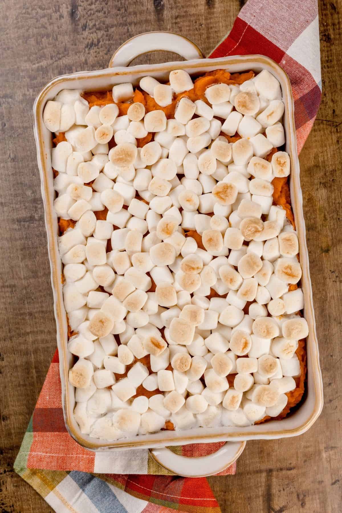 rectangle baking dish filled with sweet potatoes and covered in toasted mini marshmallows. an autumnal towel is under the dish and it is resting on a wood table.