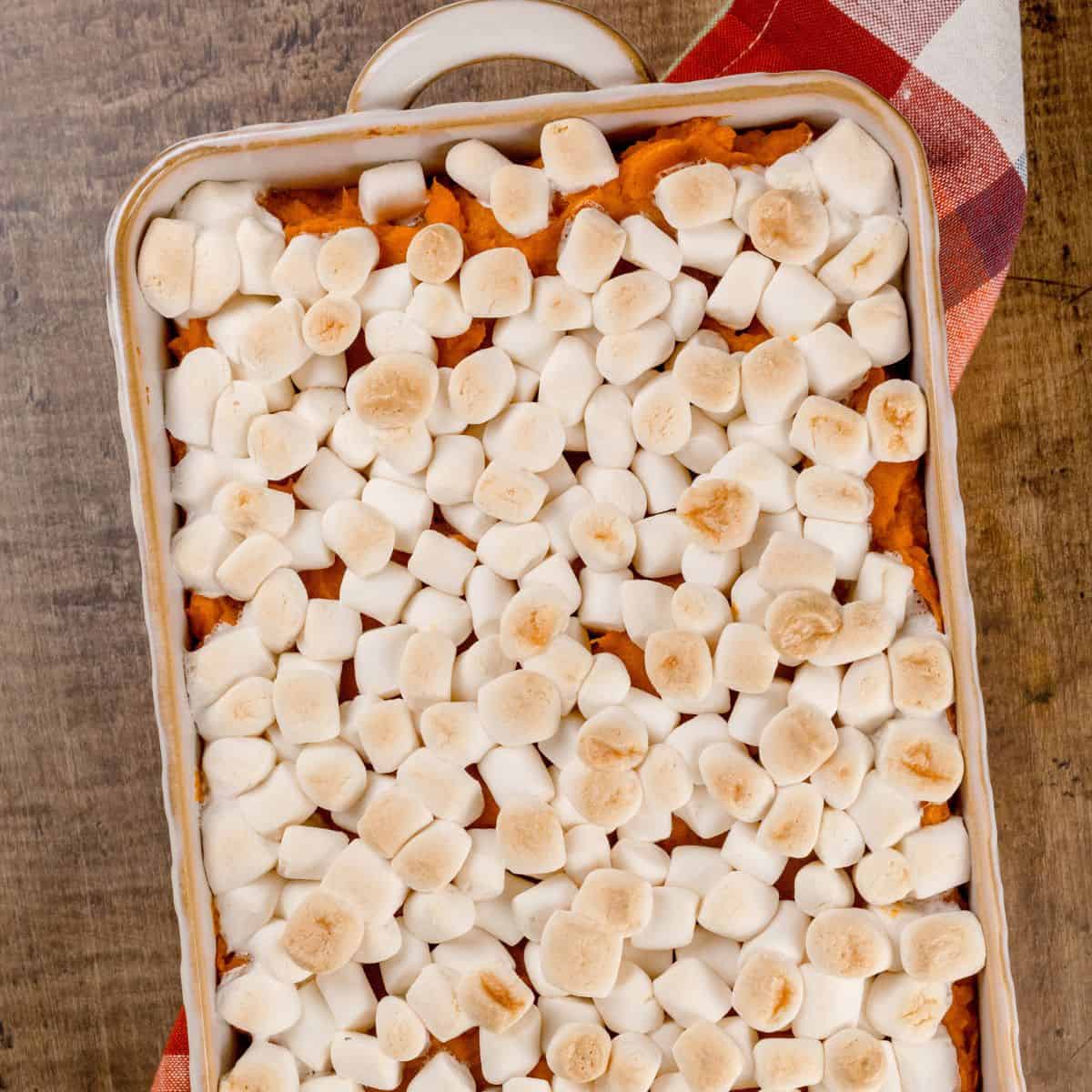 baking dish filled with roasted sweet potatoes with marshmallows resting on an autumnal kitchen towel on the wood table.