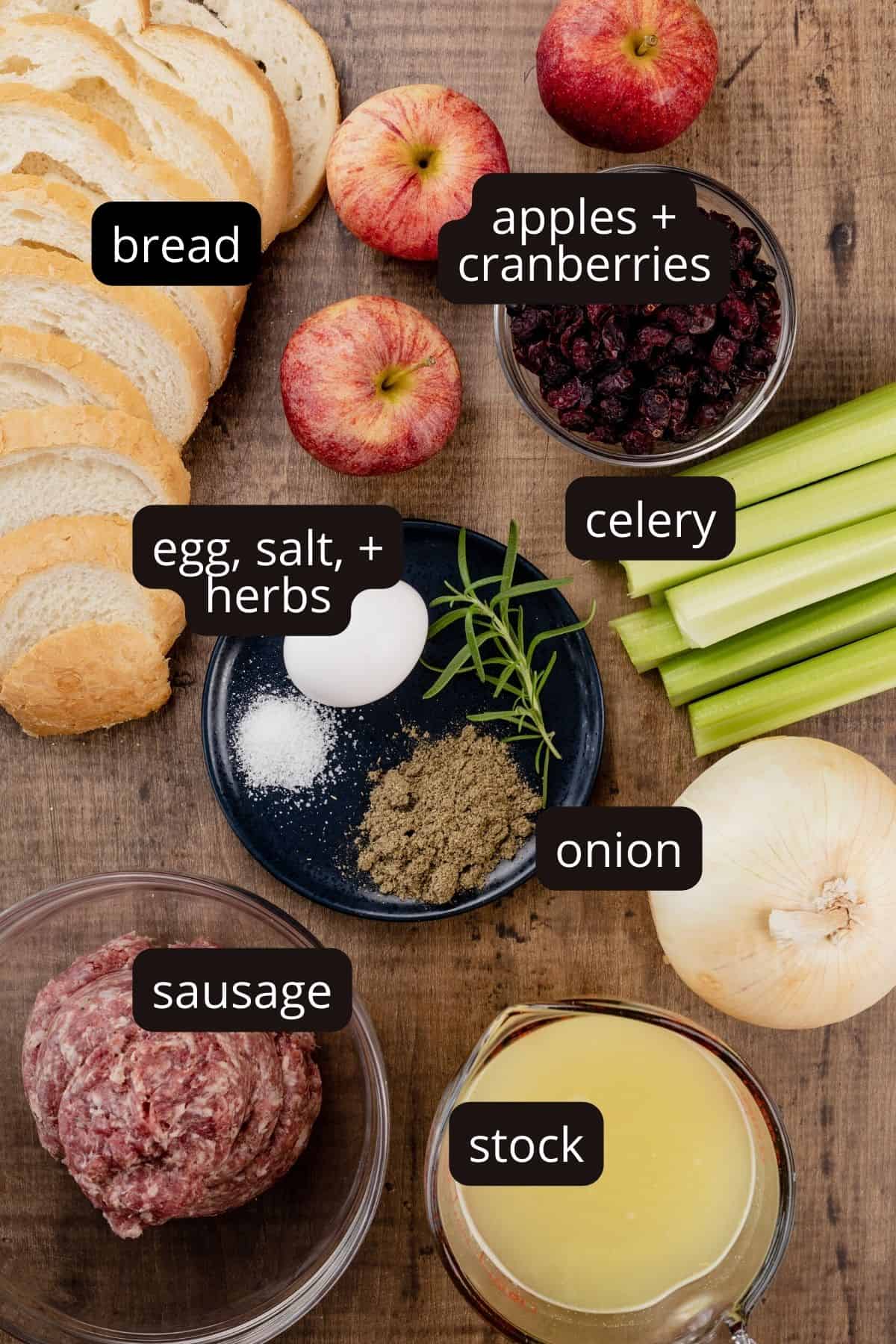 ingredients for apple sausage stuffing in various glass bowls on a wood table top. black and white labels have been added to name each ingredient.
