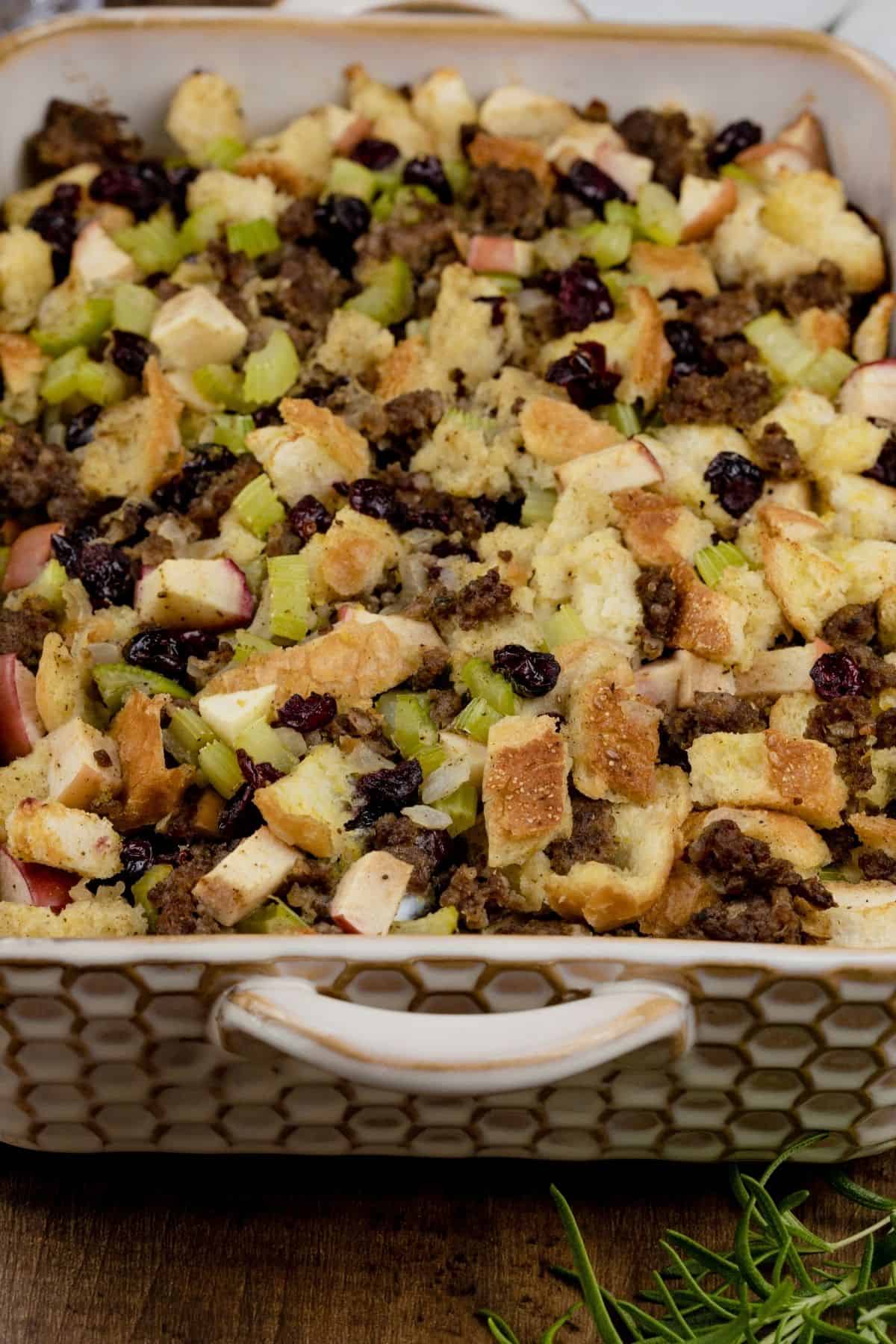 close up of apple stuffing in a honeycomb baking dish. you can see the texture of all the ingredients. a tiny sprig of rosemary in the front right corner.