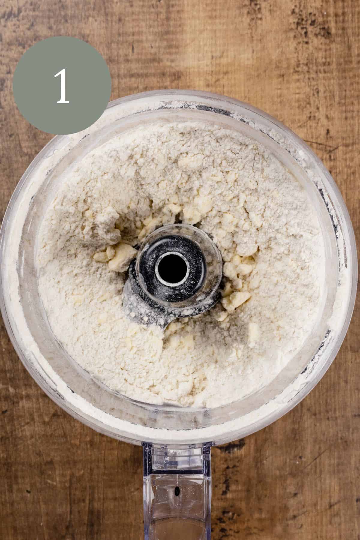 gluten free flour combined with butter in a food processor on a wood tabletop. the number 1 is in the top left corner.