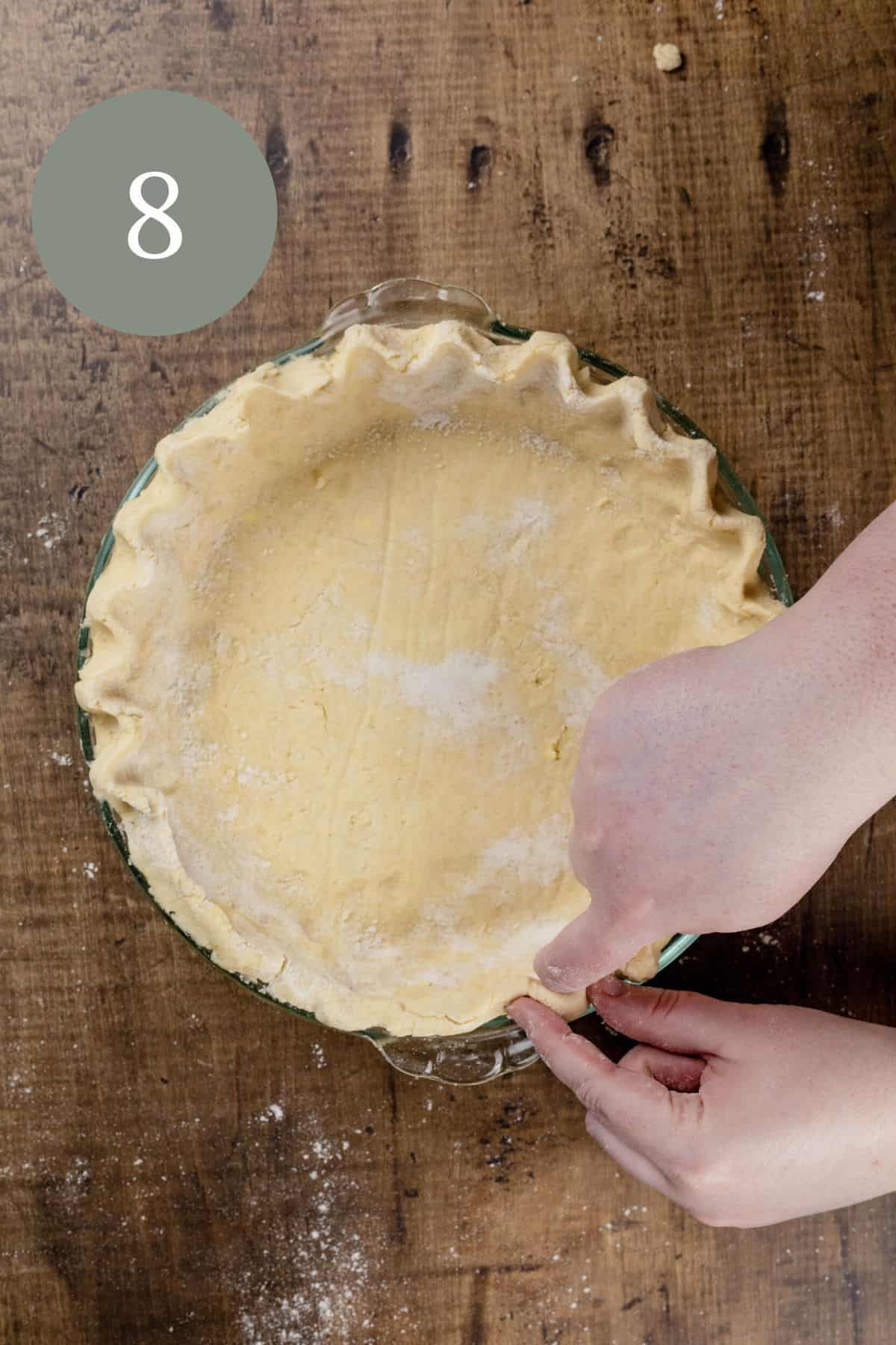 a pair of caucasian hands are crimping the edges of a gluten free pie crust on a wood tabletop. the number 8 is in the top left corner.