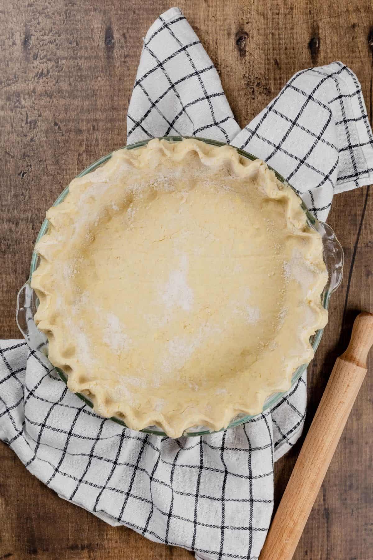 unbaked and crimped gluten free pie crust in a glass pie pan. it is resting on a blue and white towel next to a rolling pin on a wood tabletop.