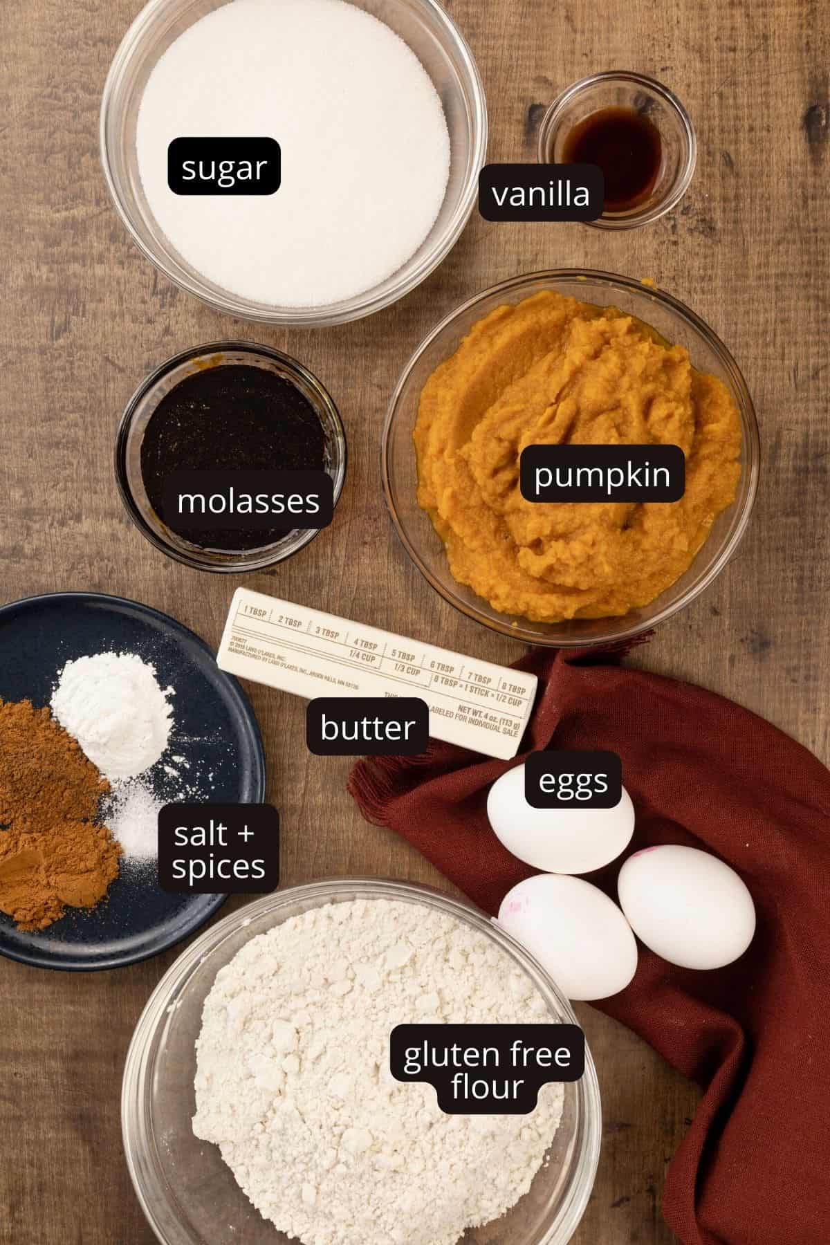 ingredients for gluten free pumpkin muffins in various glass bowls on a wood tabletop. black and white labels have been added to name each ingredient.