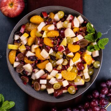 big bowl of thanksgiving fruit salad with fresh mint leaves on the side of the bowl. the bowl is resting on a dark red towel on black marble countertops. fresh fruit like apples and grapes surround the bowl.
