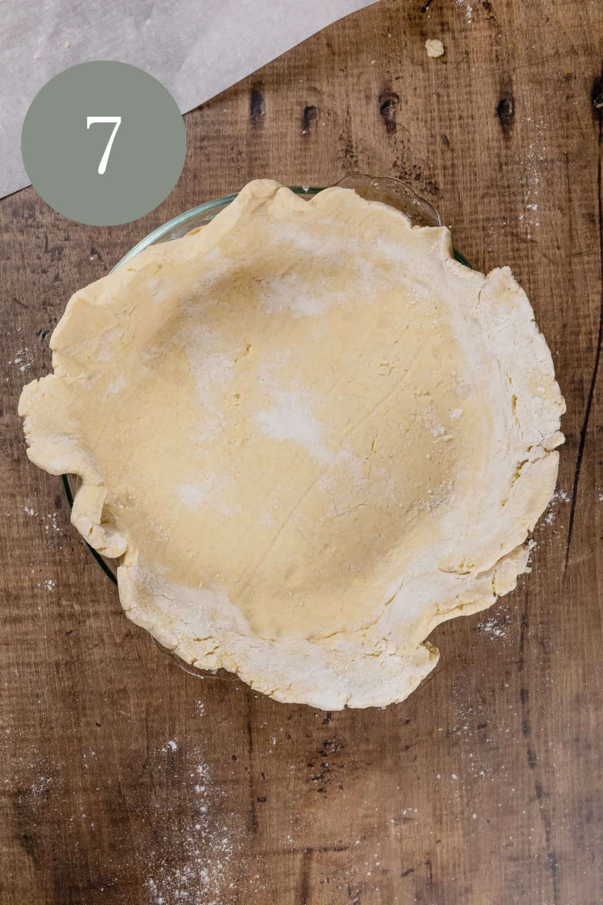 a gluten free pie crust without trimmed edges on a wood tabletop. parchment paper and extra flour are seen on the edges of the photo. the number 7 is in the top left corner.